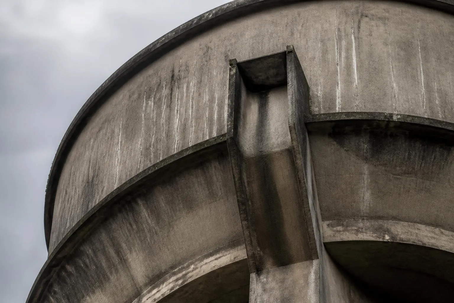 Cuve en béton d’un château d’eau vue depuis le pied de la tour, avec la goulotte de trop-plein en saillie sous la ceinture.