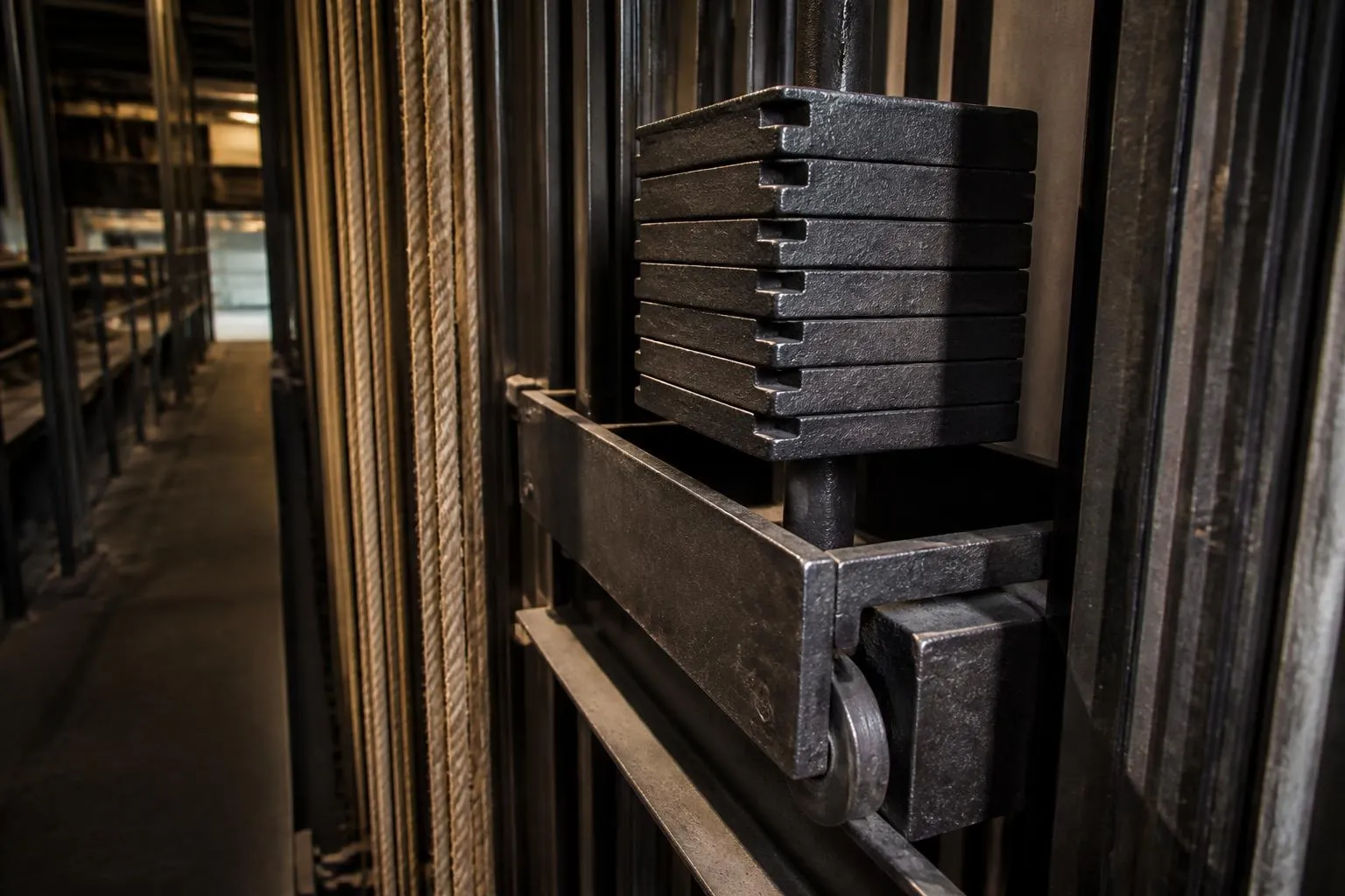 Un chariot de contrepoids chargé de pains de fonte crantés dans sa cheminée métallique, au bord d’une passerelle de théâtre